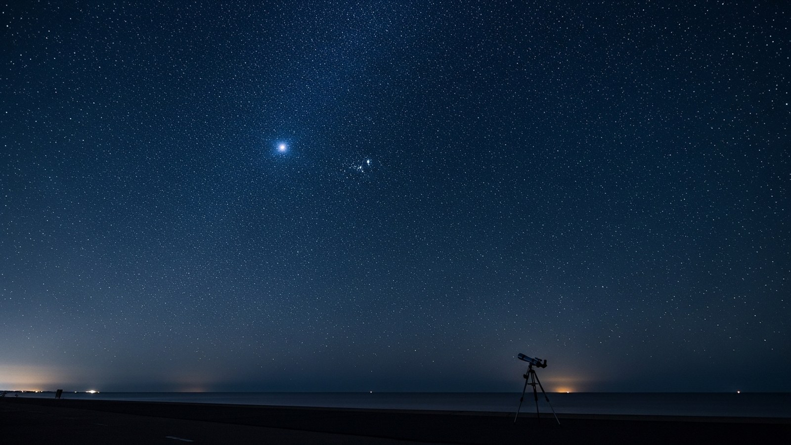 A telescope on a tripod silhouetted against a deep star field over a dark sea horizon at night